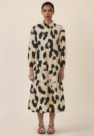 Frau trägt ein beiges Midikleid mit großen schwarzen unregelmäßigen Flecken, langen Puffärmeln, geknöpftem Kragen und braunen Riemchensandalen.