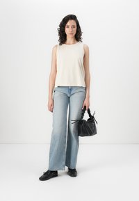 Top sin mangas color crema, jeans acampanados de un azul claro y un bolso negro con asas trenzadas. El modelo lleva zapatos negros, contra un fondo liso.