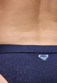 Κατώτερο μέρος της πλάτης ενός ατόμου που φοράει μπλε σκούρο ριμπ υφασμάτινα μαγιό με μικρές λευκές κουκκίδες και ένα λευκό λογότυπο σε σχήμα καρδιάς.