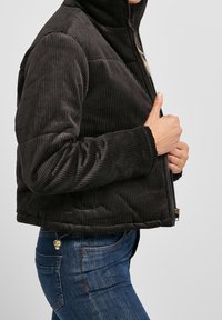 Czarna, puchowa kurtka z sztruksu z wysokim kołnierzem, zapinana na zamek, o żebrowej teksturze i bocznymi kieszeniami, noszona na niebieskich dżinsach.