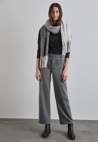 Graue weit geschnittene Hosen mit strukturiertem Finish, kombiniert mit einem schwarzen gerippten Pullover und einem grau-weißen Fransenschal, abgerundet mit schwarzen Stiefeln.