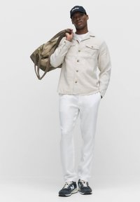 Beige linnen overhemd met knopen, witte broek en donkere sneakers. Een khaki sporttas in de hand. Eenvoudig design, neutraal kleurenpalet.