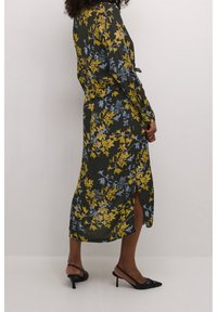 Robe maxi florale en vert foncé, ornée de motifs jaunes et bleus, manches longues et fente sur le côté. Portée avec des escarpins noirs.