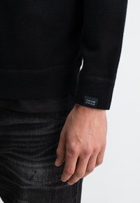 Schwarzer gestrickter Pullover mit geripptem Saum und Bündchen, mit einem Markenetikett am Bündchen. Getragen über einem schwarzen Schichthemd und grauer Jeans.