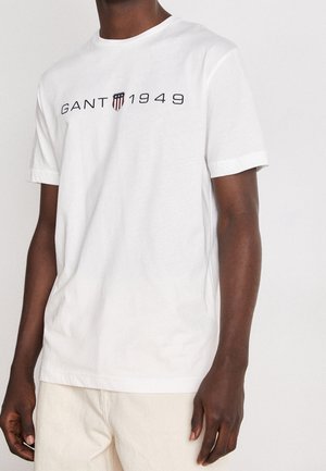 Uomo che indossa una t-shirt bianca a maniche corte con testo nero "GANT 1949" e un piccolo logo a scudo sul petto, abbinata a pantaloni chiari.