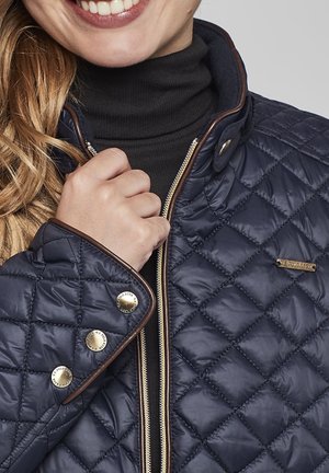 Veste matelassée bleu marine avec quincaillerie dorée, fermeture éclair et finitions marron. Comprend des boutons-pression et un col haut pour plus de chaleur.