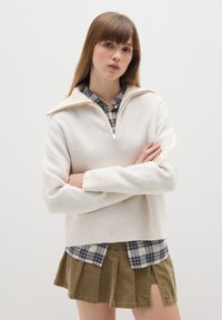 Cremfarbener Strickpullover mit geripptem Saum und halbem Reißverschluss, über ein kariertes Flanellhemd getragen, kombiniert mit einem hellbraunen Faltenrock.