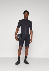 Tenue de cyclisme noire comprenant un maillot à manches courtes avec une fermeture éclair frontale et un cuissard rembourré. Le modèle tient un casque noir et porte des chaussures de cyclisme.