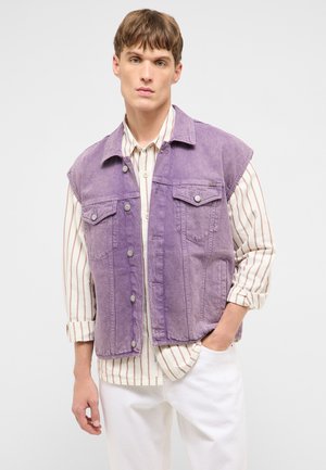 Young man wearing a sleeveless purple denim vest over a white shirt with brown vertical stripes and white pants, standing with one hand in pocket.