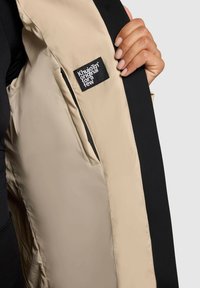Intérieur de veste beige et noire avec une texture lisse. Comprend une étiquette noire portant l'inscription "Khuiomi original pour quelques-uns." Main tenant le tissu.