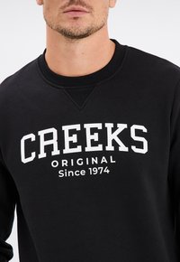 Sudadera negra con cuello redondo, puños acanalados y "CREEKS ORIGINAL Desde 1974" en texto blanco en la parte delantera. Material suave de mezcla de algodón.
