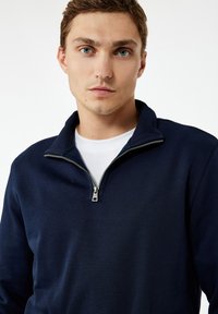 Granatowy pulower z suwakiem na ćwierć, wykonany z miękkiego materiału, z wysokim kołnierzem i srebrnym zamkiem. Nałożony na biały T-shirt pod spodem.