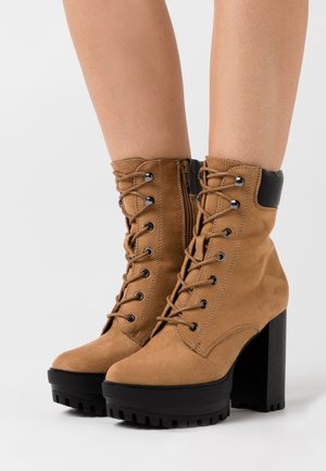 Brown suede lace-up boots with black platform chunky heels and side zippers worn on feet against a plain background.