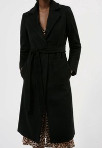 Manteau noir en tissu texturé, avec une taille à nouer, un col et deux poches avant. Porté par-dessus une robe à imprimé léopard.