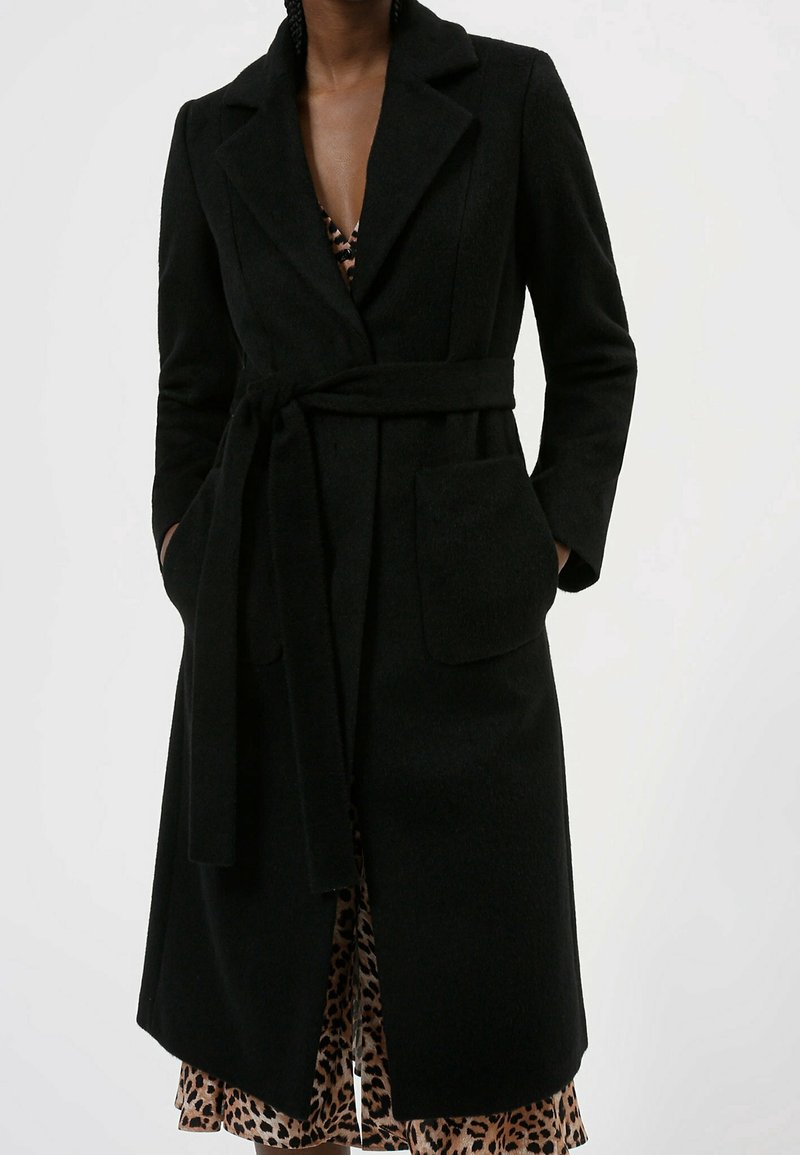 Manteau noir en tissu texturé, avec une taille à nouer, un col et deux poches avant. Porté par-dessus une robe à imprimé léopard.