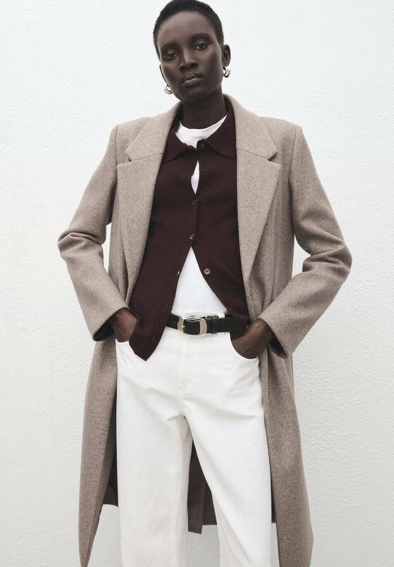 Cappotto di lana beige sopra una camicia bordeaux con bottoni, t-shirt bianca e jeans bianchi, cinchati con una cintura nera con fibbia argentata.