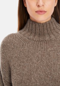Beige gestrickter Pullover mit hohem Rippenkragen. Die Textur ist weich und dick und weist ein subtil verteiltes Muster auf.