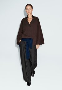 Vrouw in een losse donkerbruine knoopshirt, wijde grijze broek, zwarte schoenen en een marineblauwe trui om haar middel, staande tegen een witte muur.