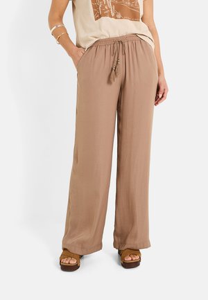 Pantalones ligeros de color marrón claro, holgados y con cintura ajustable mediante cordón, decorados con borlas, combinados con una camiseta gráfica beige y sandalias de plataforma marrones.