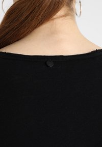 Maglietta nera con scollatura rotonda, caratterizzata da un orlo grezzo e un piccolo bottone nero sul retro. Il tessuto sembra morbido e liscio.
