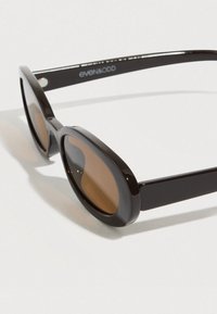 Μαύρα γυαλιά ηλίου με οβάλ φακούς και καφέ απόχρωση. Λεπτό πλαστικό πλαίσιο με γυαλιστερό φινίρισμα και ελάχιστες σχεδιαστικές λεπτομέρειες.