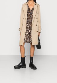 Trinchera beige con botones dorados sobre un vestido negro con un patrón abstracto en naranja y crema. Incluye botas negras hasta el tobillo y un bolso.