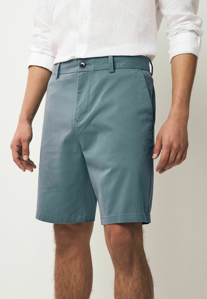 Shorts pentru bărbați dintr-un amestec de bumbac de culoare teal, cu un front plat, bucle pentru curea, buzunare laterale și un închidere cu un singur buton. Textură netedă și lungime până la genunchi.