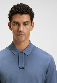 Ung man med kort, lockigt hår som bär en blå polo med två knappar och "BOSS"-logotypen broderad under knäppningen.