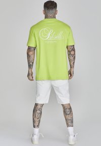 T-shirt di cotone verde brillante con maniche corte, caratterizzato da un logo bianco sul retro. Indossato con pantaloni corti di jeans bianchi e sneakers.