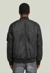 Giacca bomber nera con texture liscia, polsini e orlo a coste, design imbottito con dettagli a cuciture lungo la schiena.