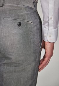 Pantaloni gri, ajustați, cu o textură netedă, având un sistem de închidere cu buton la un buzunar din spate și o talie adaptată. Mână într-o cămașă albă. S