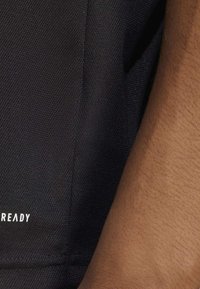 Tessuto nero con texture e il testo "READY" in bianco nella parte inferiore. Mostra uno sfondo scuro e un braccio di carnagione chiara parzialmente visibile.