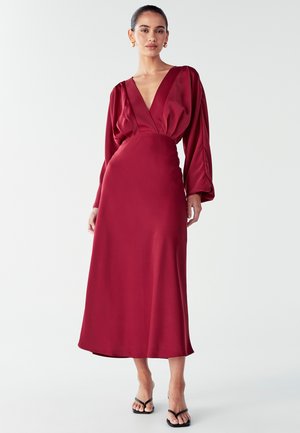 Raudonas satino suknelė su giliu V formos iškirpimu, ilgomis pūkuotomis rankovėmis ir prigludusia juosmens linija, besileidžiančia iki vidurio blauzdų. Dėvima su juodais sandalais.