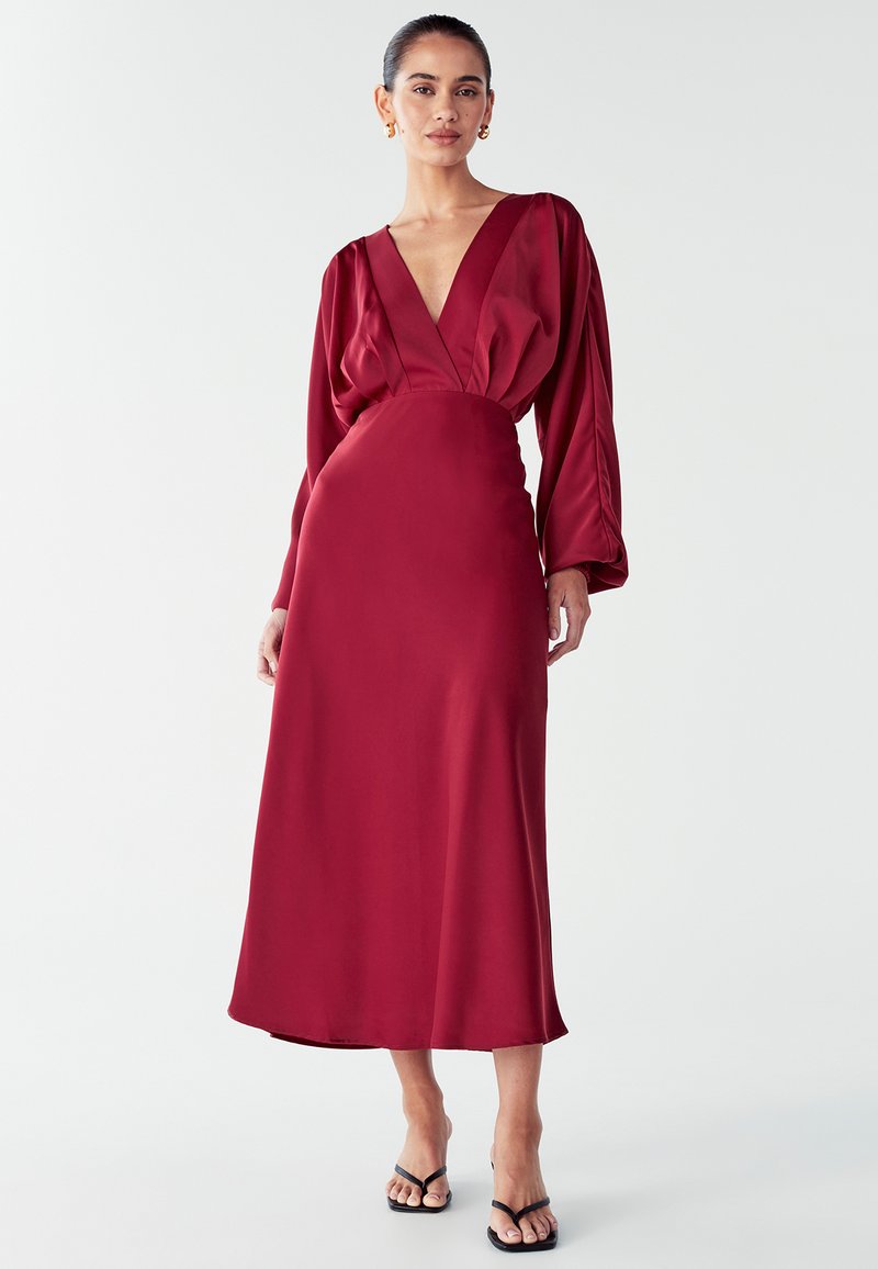 Raudonas satino suknelė su giliu V formos iškirpimu, ilgomis pūkuotomis rankovėmis ir prigludusia juosmens linija, besileidžiančia iki vidurio blauzdų. Dėvima su juodais sandalais.