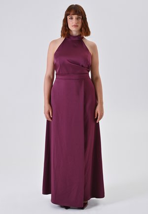 Frau mit schulterlangem Haar, die ein ärmelloses, hochgeschlossenes, bodenlanges, pflaumenlila Kleid vor einem schlichten, hellen Hintergrund trägt.