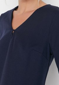 Haut bleu marine avec un décolleté en V, présentant un petit détail découpé et une poche sur le côté gauche. Le tissu est lisse et léger.