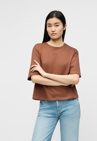 Γυναίκα με μακριά μαύρα μαλλιά που φοράει καφέ t-shirt EA7 με κοντά μανίκια και ανοιχτόχρωμο τζιν, στέκεται με τα χέρια σταυρωμένα μπροστά σε απλό φόντο.