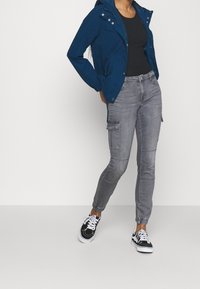 Kobieta w niebieskiej kurtce z kapturem, czarnej bluzce, szarych spodniach cargo i czarno-białych sneakersach stoi na jednolitym tle.