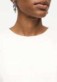 Κοντινό πλάνο του λαιμού και των χειλιών ενός ατόμου που φοράει ασημένια σκουλαρίκια κρίκους με συνδέσμους αλυσίδας και λευκό μπλουζάκι με ραφή λαιμού, πάνω σε λευκό φόντο.