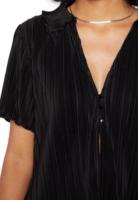 Zwarte geplooide blouse met een kraag, korte mouwen en knoopsluiting, met een diepe V-hals en een gladde textuur. Zilveren choker zichtbaar.