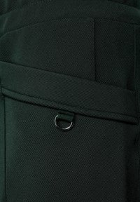 Mörkgrön tyg med en texturerad yta, med en frontficka och en metallring som accent. Designen inkluderar synliga sömnadsdetaljer.