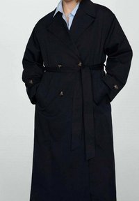 Trench-coat bleu foncé à double boutonnage, avec deux poches frontales, une ceinture et des boutons dorés. Confectionné en tissu robuste.