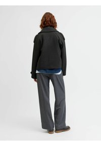 Črn teksturiran jopič z širokim ovratnikom in ramenskimi epauletti, prelit preko modre jeans srajce, v kombinaciji s sivimi širokimi hlačami.