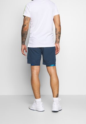 Mann steht mit dem Rücken zur Kamera, trägt ein weißes T-Shirt, marineblaue Shorts über blauen Kompressionsleggings, weiße Socken und weiße Turnschuhe.