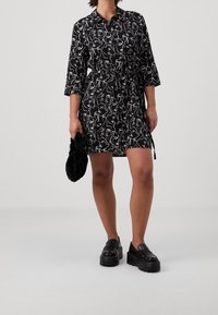Abito camicia nero con motivi astratti bianchi, chiusura con bottoni frontale, vita cinchata, maniche a tre quarti, abbinato a mocassini neri massicci.