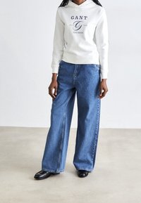 Bílá mikina s kapucí a modrým logem 'GANT', modré džíny se širokým střihem, světlá modrá denimová textilie, černé boty s špičatou špičkou.