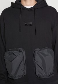 Svart adidas hoodie med dragsko som har två stora rektangulära framfickor med dragkedja och en liten adidas-logotyp på bröstet.