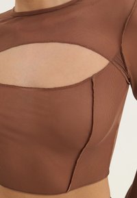 Brun kort topp i texturerat tyg. Har rund halsringning och utskärning på bröstet. Sömmarna är synliga längs kanterna.