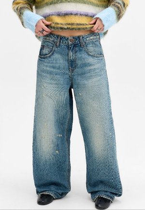 Personne portant un jean bleu délavé à jambes larges, un pull rayé et duveteux, et des chaussures noires à bouts pointus, tenant l'ourlet du pull.