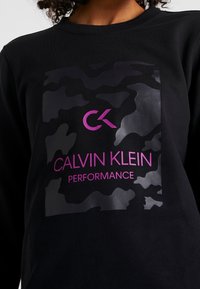 Mustat collegepaidat, joissa on tummia sävyjä oleva camotyyppinen kuvio. Sisältää kiiltävän vaaleanpunaisen logon ja tekstin "CALVIN KLEIN PERFORMANCE."